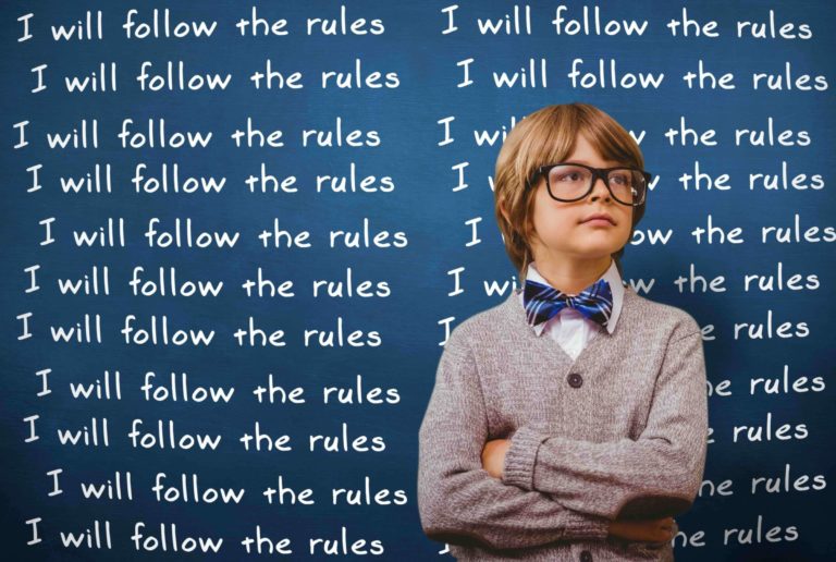 Young boy standing in front of a chalkboard reading ‘I will follow the rules’.