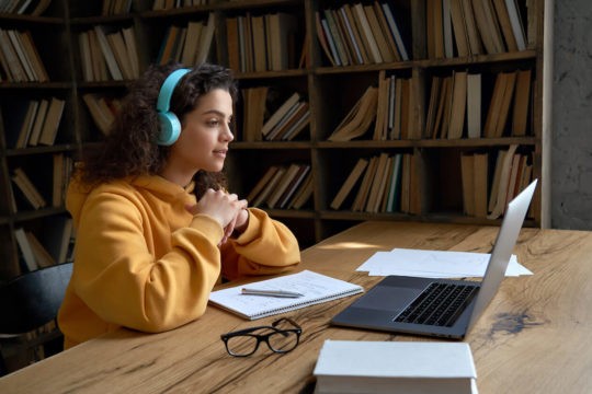 Girl with headphones on watching a Zoom class while taking notes.