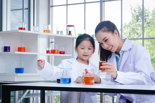 Female teacher and child using look a measuring cups filled with colored liquid