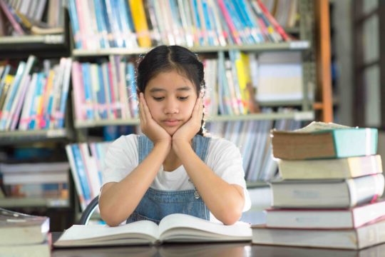 Young girl sitting at a library table with books looking bored with her hands on her face