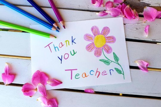 A piece of paper says “thank you teacher” with a drawing of a flower and surrounded by colored pencils used to create the thank you note.