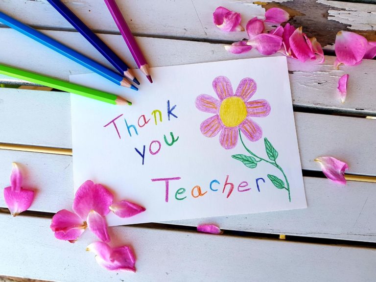 A piece of paper says “thank you teacher” with a drawing of a flower and surrounded by colored pencils used to create the thank you note.