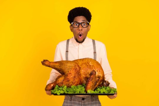 A man in glasses holds a Thanksgiving turkey with a shocked look on his face, in front of an isolated yellow background.