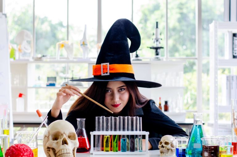 A girl in a witch costume sits in a science lab surrounded by lab experiment equipment.