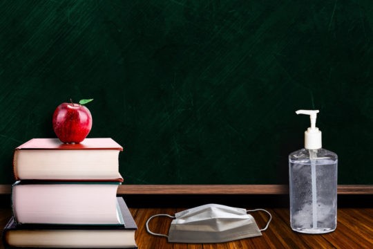 A stack of textbooks with an apple on top next to a face mask and hand sanitizer