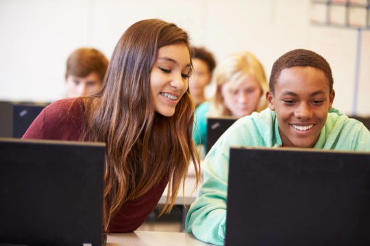 Two smiling high school students on computers doing research