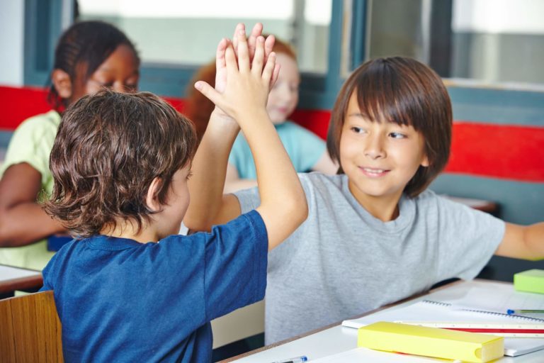 Two young students sitting at their desks high-fiving each other.