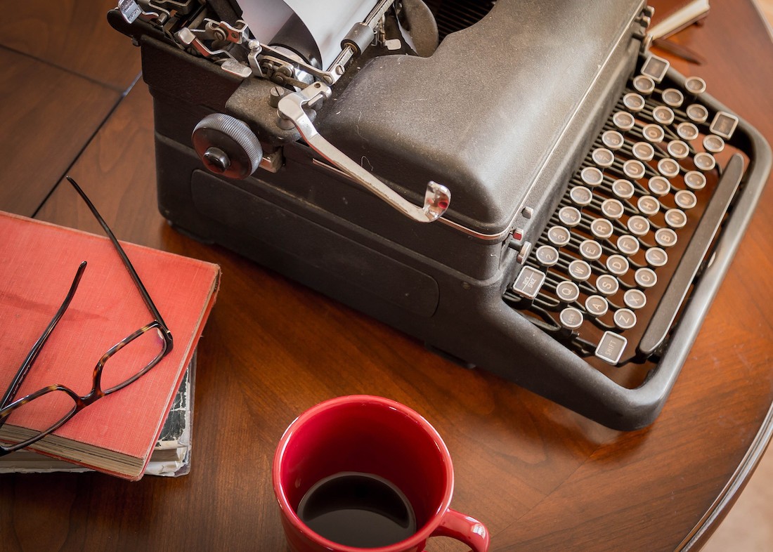 Black typewriter sitting on a table with books, glasses, and coffee